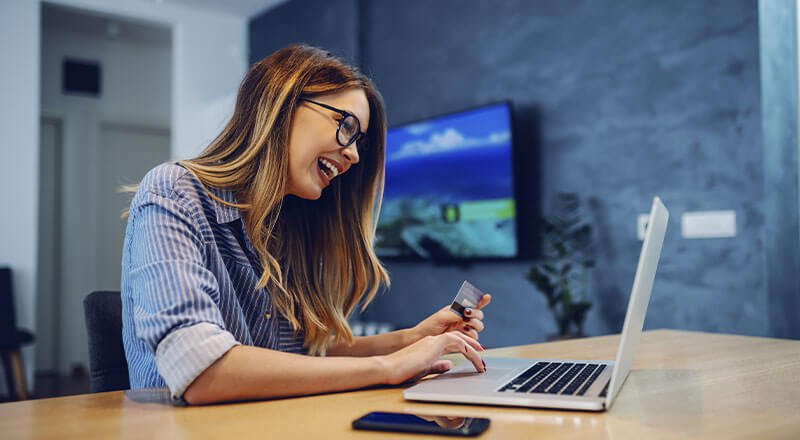 Uma mulher branca de óculos sorrindo enquanto olha a tela de um notebook cinza e toca nos teclados do mesmo.