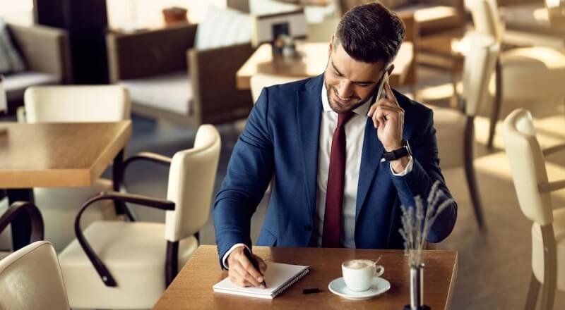 Um homem de terno azul sentado em uma mesa falando ao celular enquanto anota algo em um caderno. Há também uma xícara de café branca na mesa.